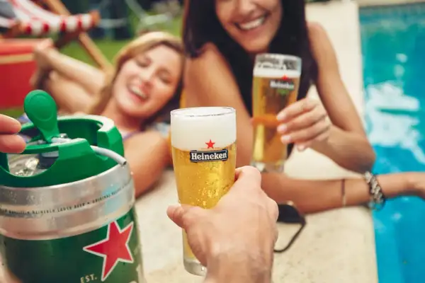 Un fusto di birra Heineken pronto per essere spillato, con due donne sorridenti che brindano con bicchieri di birra vicino a una piscina.
