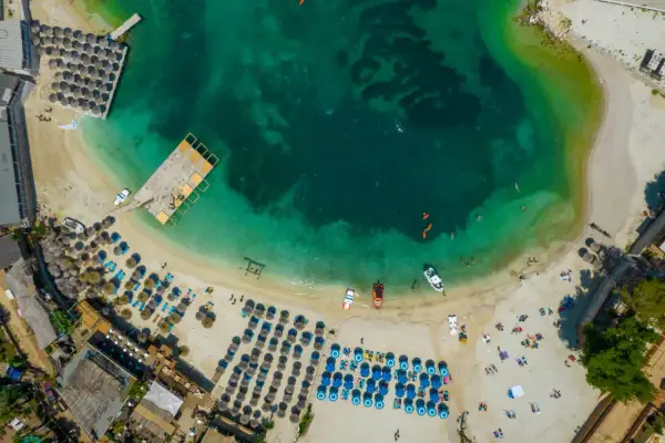 Widok z lotu ptaka na turkusowe morze i piaszczystą plażę z rzędami leżaków i parasoli. Woda jest krystalicznie czysta, widać ludzi pływających i łodzie. To idealne miejsce na relaks na **tirana plaże**.
