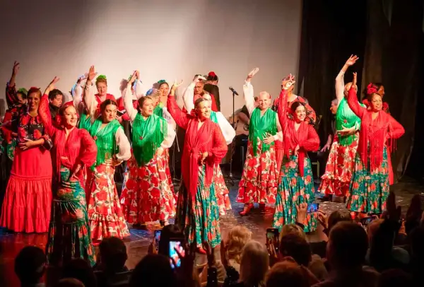 Un grupo de mujeres baila sevillanas de amor con trajes coloridos. Sus faldas amplias y mantones vibrantes llenan el escenario de alegría.