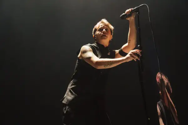Lead singer of Nothing But Thieves, Conor Mason, belts into a microphone on a dark stage, his arm tattoos visible.