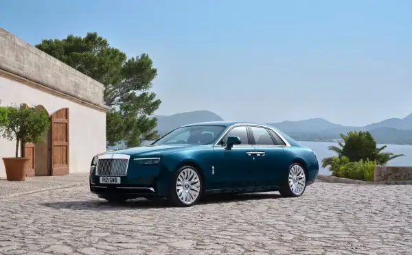 Elegancki Rolls-Royce Ghost w kolorze głębokiego turkusu zaparkowany na kamiennym dziedzińcu, z widokiem na morze i góry.