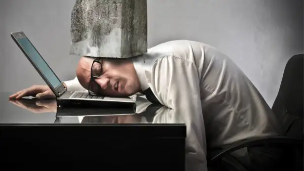 Man med sten på huvudet, utmattad vid sin laptop. Symboliserar digital stress och överväldigande arbetsbelastning.