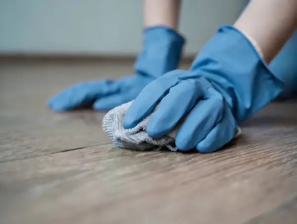 Mani con guanti blu puliscono un pavimento in legno con un panno, creando un detersivo pavimenti fai da te.