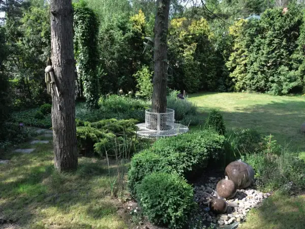 Urokliwy ogród w lesie z kamiennymi kulami tworzącymi wodną kaskadę, ozdobną ławką wokół drzewa i bujną zielenią.