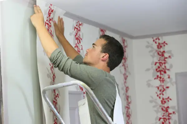 Mann auf Leiter tapeziert Wand mit rotem Blumenmuster. So tapeziert man Schritt für Schritt.