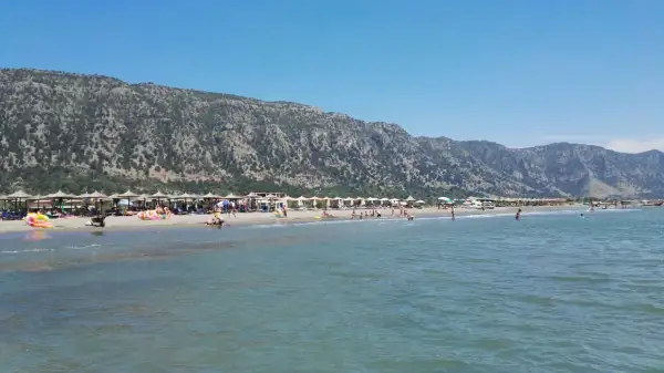 Słoneczny dzień na plaży Velipoja. Ludzie odpoczywają pod parasolami, kąpią się w morzu, a w tle wznoszą się górzyste tereny.