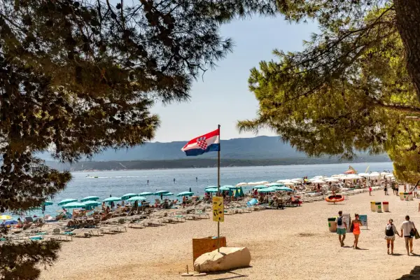 Chorwacka flaga powiewa nad pełną plażą z leżakami i parasolami. W tle błękitne morze i góry.