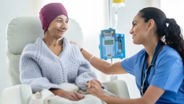 Eine Krankenschwester tröstet eine Patientin während des Chemotherapieablaufs. Die Patientin trägt eine lila Kopfbedeckung und eine Strickjacke.