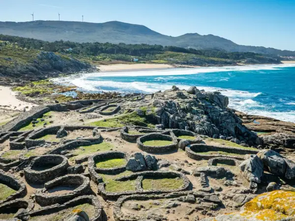 Ruinas circulares de piedra del castro de Baroña, con el mar azul y una playa de arena al fondo.