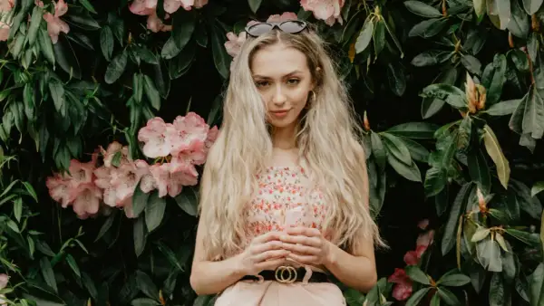 Młoda kobieta o blond włosach i słowiańskiej urodzie, w kwiecistej sukience, stoi na tle bujnej zieleni i różowych kwiatów.