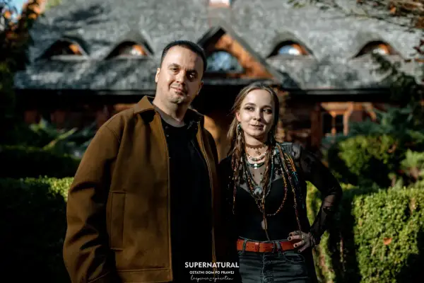 Para pozuje przed domem z kamiennym dachem. Mężczyzna w brązowej kurtce i kobieta w czarnej bluzce z wieloma naszyjnikami. Wyglądają jakby wyszli z planu filmu o straszydłach.