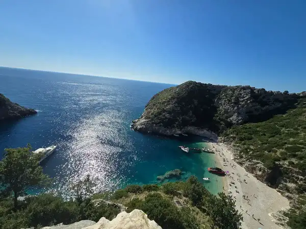 Słoneczna zatoka grama z turkusową wodą, kamienistą plażą pełną ludzi i łodziami przycumowanymi do brzegu.
