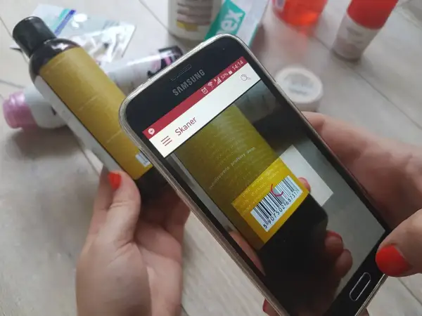 Ręce trzymają smartfon z otwartą aplikacją do sprawdzania składu kosmetyków. Na ekranie widoczny jest skaner kodu kreskowego skierowany na etykietę butelki.