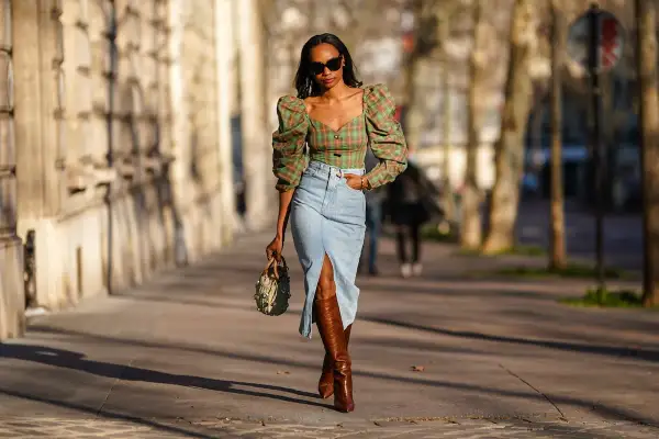 Mujer con top de cuadros y falda vaquera larga con abertura, botas altas y bolso.