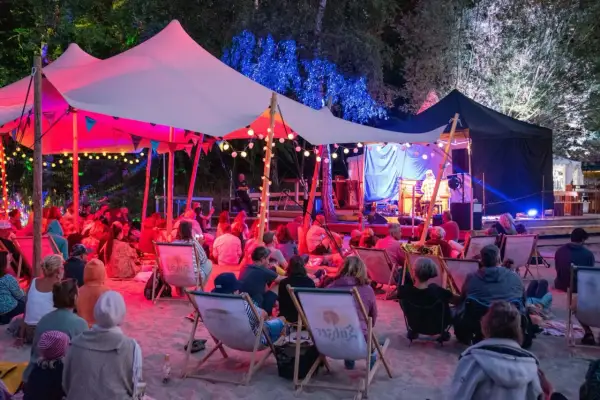 Menschen genießen ein **musik und stille festival** unter einem beleuchteten Zelt. Musiker spielen auf einer Bühne, während das Publikum auf Liegestühlen im Sand sitzt.