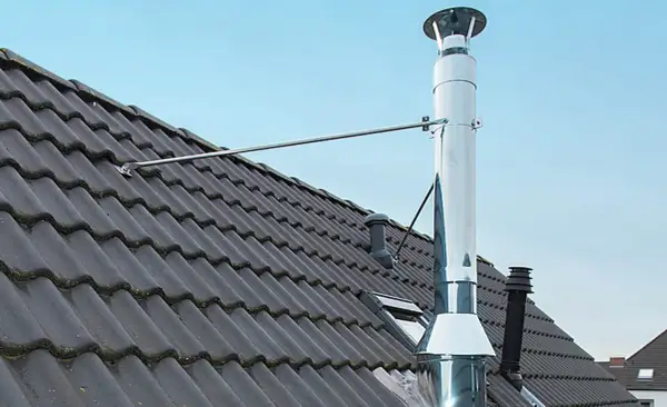 Edelstahl-Ofenrohre verbinden sich auf einem dunklen Ziegeldach. Ein Metallarm stützt das glänzende Rohr.