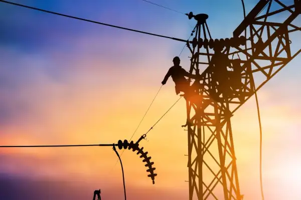 Sylwetka pracownika na tle zachodzącego słońca, naprawiającego linię energetyczną. Widać izolatory i konstrukcję wieży, a także potencjalny łuk elektryczny.