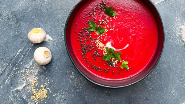 Eine leuchtend rote Bete Suppe, garniert mit Sesam und Petersilie, neben zwei Knoblauchzehen.