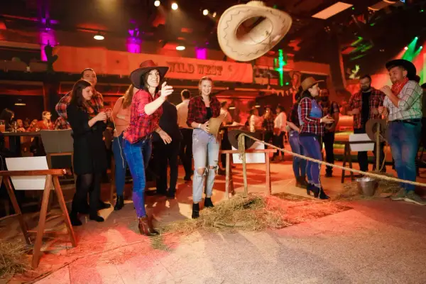 Dziewczyny w kapeluszach i kratkach bawią się na imprezie w stylu country. Rzucają kapeluszem, a w tle widać napis "Wild West".