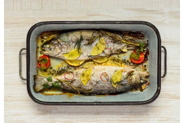 Deux truites au four, garnies de rondelles de citron, d'oignon rouge, de tomates cerises et d'herbes aromatiques, dans un plat en émail.