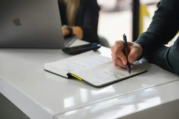 Dłoń pisząca długopisem w notatniku na białym biurku, obok laptopa.