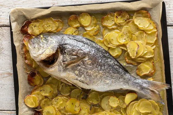 Orata al forno dorata e succulenta, adagiata su un letto di patate affettate e cotte a puntino, profumate di erbe aromatiche.