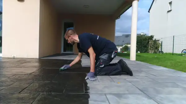 Un homme agenouillé effectue le nettoyage de pierre naturelle extérieur avec une éponge, redonnant son éclat à la terrasse.