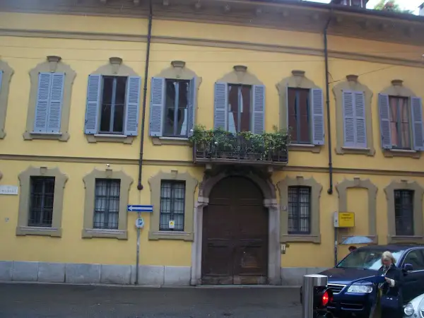 Facciata gialla del **Palazzo Belgioioso** con finestre dalle persiane azzurre e un balcone fiorito.