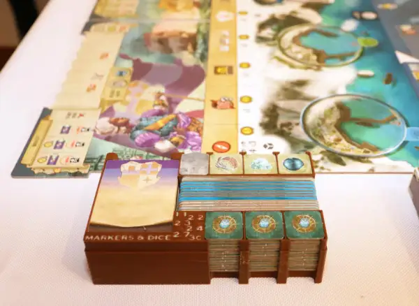 Spielkomponenten für das Dead Reckoning Brettspiel, darunter Karten, Marker und Würfel, sind ordentlich in braunen Halterungen gestapelt.