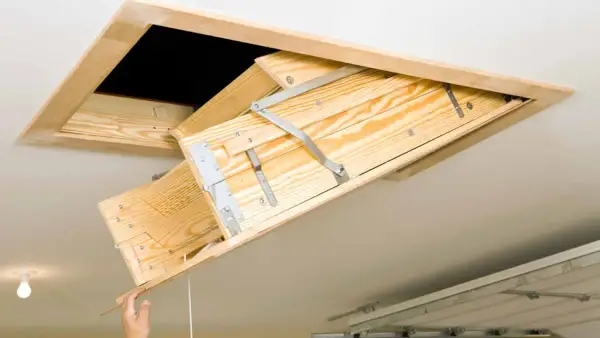 A wooden attic ladder is being installed, with a hand pulling down the first section.
