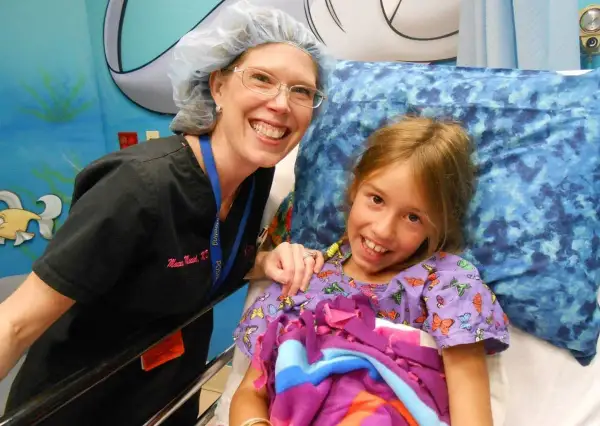 Niña sonriente en cama de hospital, junto a enfermera. Ella tiene síndrome de Marfan y recibe cuidados.