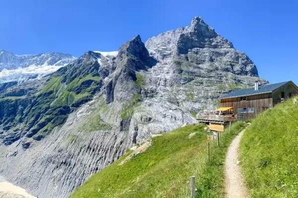 Ein Weg führt zu einer Berghütte mit Terrasse vor imposanter Schweizer Bergkulisse. Perfekt für eine Pause beim **Schweiz wandern**.