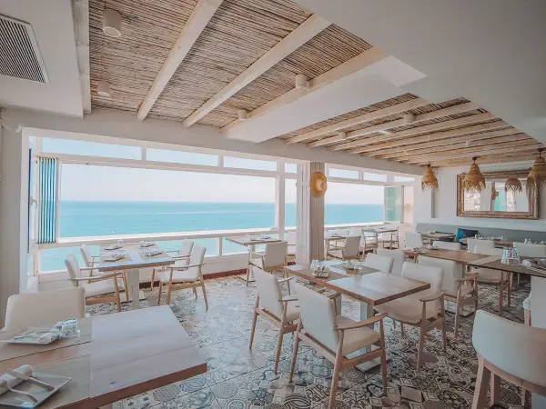 Comedor de la casa Pepe San José con vistas al mar. Mesas puestas, sillas blancas y techo de caña.