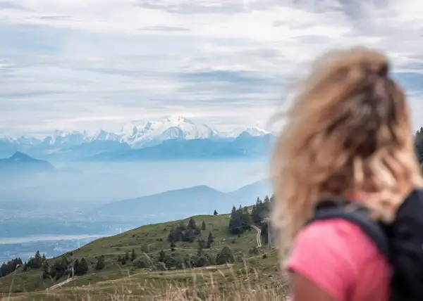 Vue panoramique sur les Alpes enneigées, avec une femme blonde en premier plan. Un sentier serpente sur la colline verdoyante, invitant au trek dans le Jura.