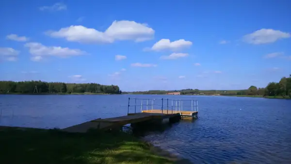 Drewniana przystań na spokojnym zalewie Drzewica. Błękitne niebo z białymi chmurami nad taflą wody i zielonymi lasami w tle.