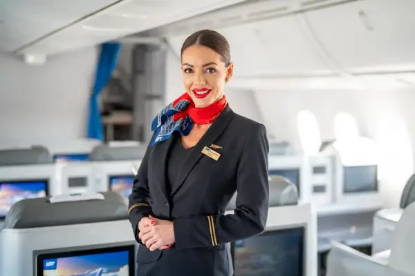 Uśmiechnięta członkini **cabin crew** w granatowym mundurze z czerwonym szalem, stoi na tle rzędów foteli w samolocie.
