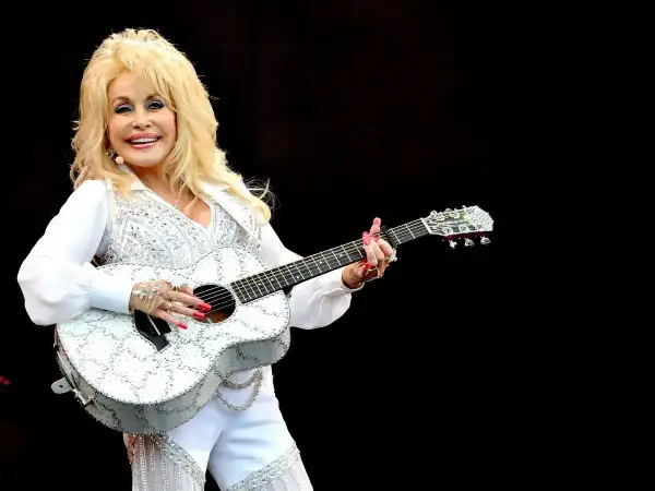 Dolly Parton, w białym, błyszczącym stroju, gra na gitarze. Wyobraź sobie, że śpiewa "Jolene", a tekst piosenki rozbrzmiewa w powietrzu.