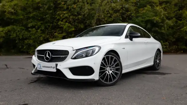 Biały Mercedes C43 AMG coupe z czarnymi felgami, zaparkowany na tle zieleni.