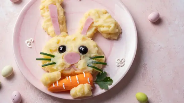 Un adorable lapin en purée de pommes de terre, parfait pour une entrée de Pâques sans œufs. Il tient une carotte et des œufs de Pâques sont autour.