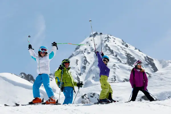 Famille en tenue de ski, bras levés, profitant du sport alpin sur une pente enneigée avec des montagnes majestueuses en arrière-plan.