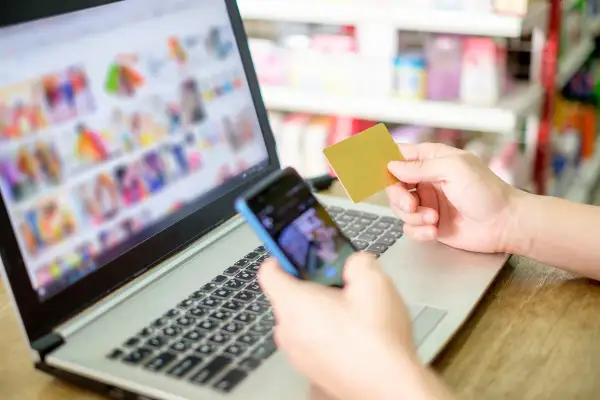 Dłoń trzymająca kartę płatniczą i telefon nad klawiaturą laptopa.