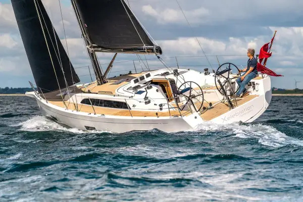 Un uomo guida una barca a vela bianca con vele nere, solcando le onde. Il nome "Xelebration" è visibile sullo scafo.
