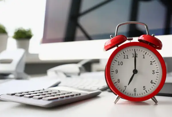 Sulla scrivania, un orologio rosso segna le 7. La calcolatrice e il computer suggeriscono che il giorno di riposo viene pagato, ma il lavoro attende.