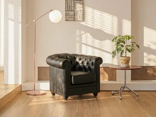 En svart chesterfieldfåtölj står bredvid ett runt sidobord med en grön växt. En kopparfärgad golvlampa ger ett mjukt ljus.