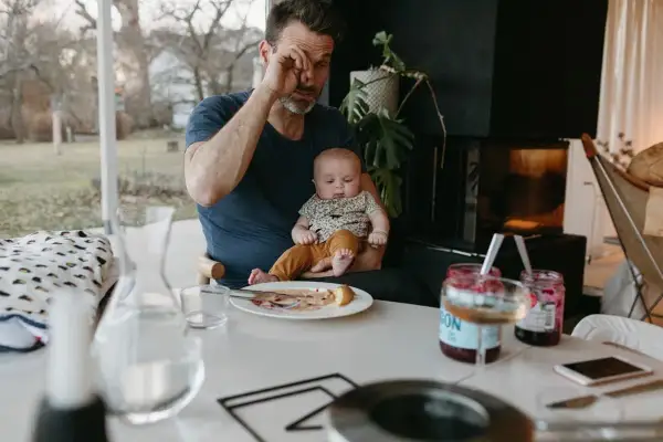 Trött pappa försöker äta frukost medan en bebis vaknar tidigt. Barnet sitter i pappans knä, en tallrik med mat står på bordet.