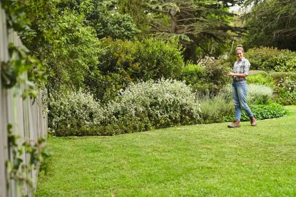 Mujer sonríe en un jardín exuberante, disfrutando del cuidado del césped en primavera. El césped verde y los arbustos floridos crean un paisaje idílico.