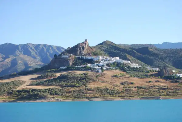 Pueblo blanco en la cima de una colina rocosa, con un castillo en ruinas. Un lugar ideal que ver cerca de Ronda, con un embalse azul en primer plano.