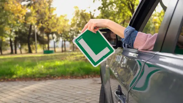 Mano sacando una L verde de autoescuela por la ventana de un coche. ¡Ya estoy lista para apuntarme a la autoescuela!