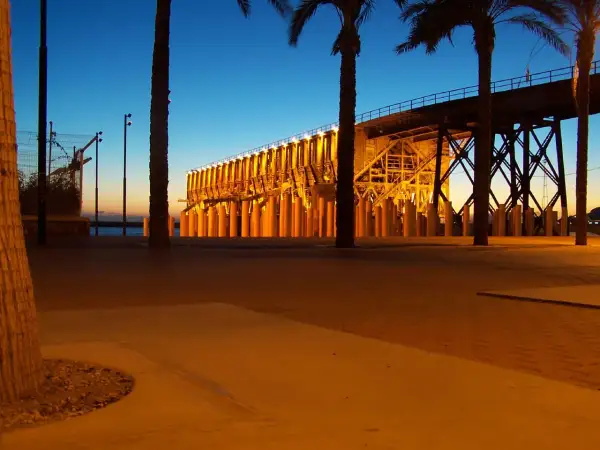 Atardecer en el parque de las almadrabillas, con palmeras y un puente iluminado sobre el mar.