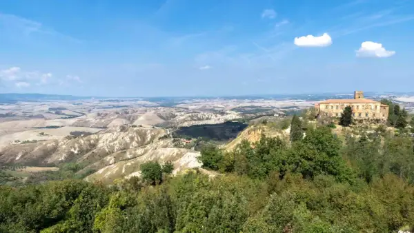 Panoramica delle **balze di Volterra**, con un casale in pietra in primo piano e dolci colline ondulate all'orizzonte sotto un cielo azzurro.